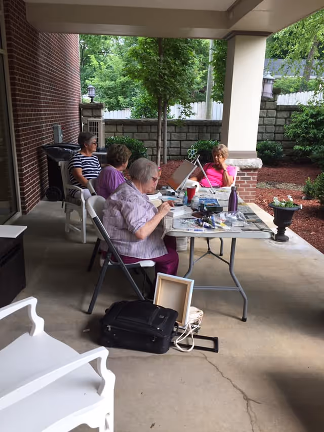 Four elderly women sitting around a table on a covered patio engaged in painting and art activities. The patio is adjacent to a brick building and overlooks a garden area with trees, shrubs, and a stone wall.
