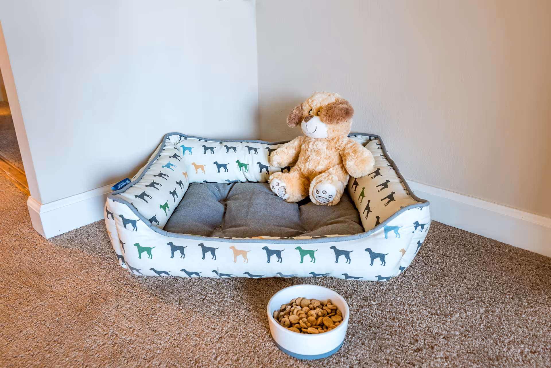 A dog bed with a plush teddy bear sits in a carpeted corner next to a bowl of kibble.