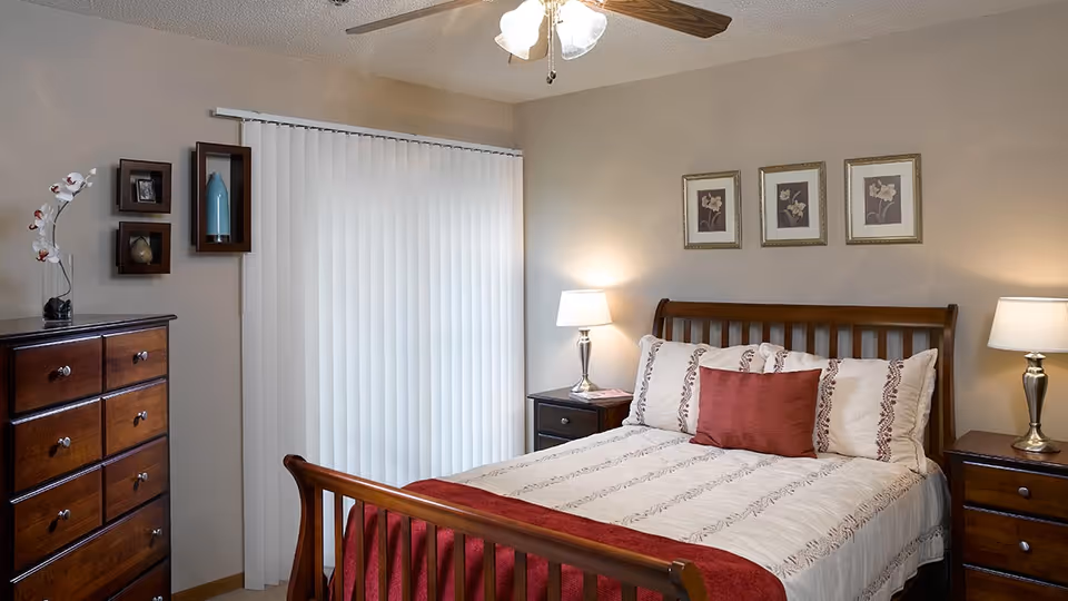 A neatly made bedroom with a wooden bed, matching dressers and nightstands, two table lamps, framed wall art, and vertical blinds covering a sliding door.