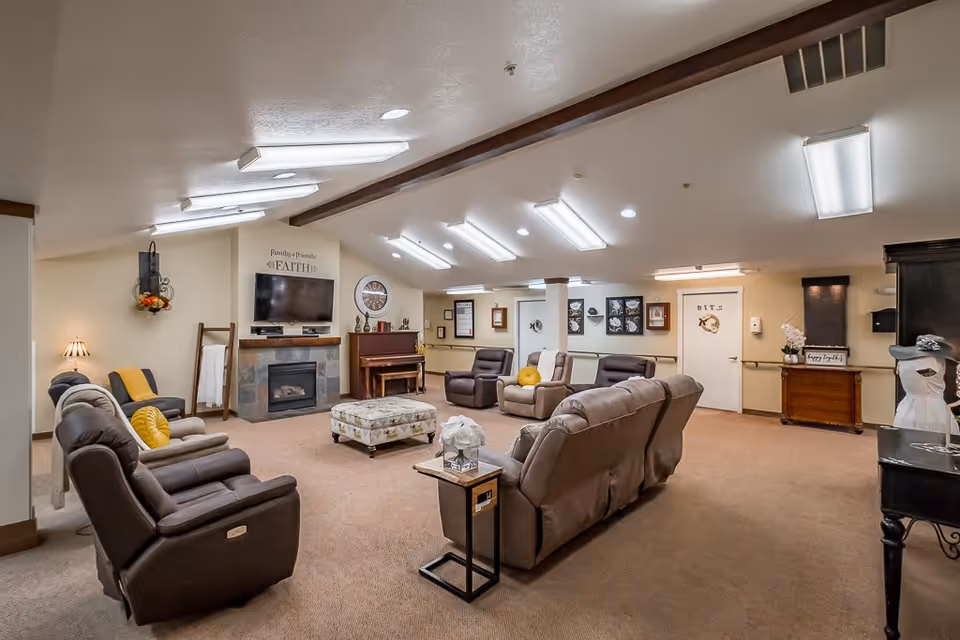 A spacious, well-lit common living area with multiple comfortable recliner chairs and sofas arranged around a central ottoman. The room features a fireplace with a mounted TV above it, a piano, wall decorations, and soft beige carpeting. The ceiling has several fluorescent light fixtures, and there are decorative items like a flower arrangement and framed pictures on the walls.