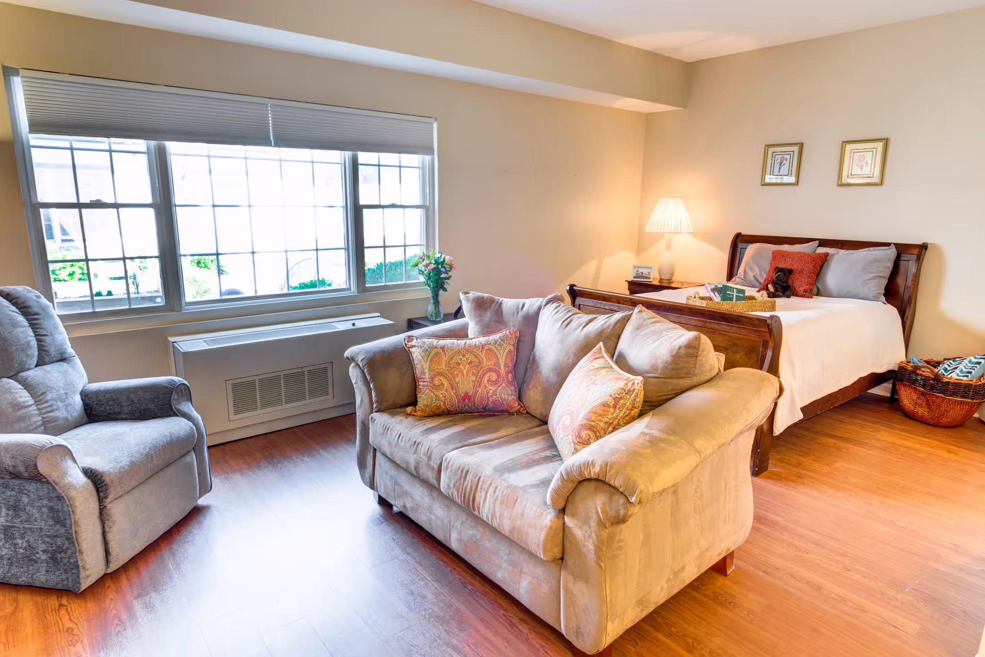 A cozy room with a large window letting in natural light, featuring a beige loveseat with patterned cushions, a gray recliner chair, and a wooden bed with white bedding and decorative pillows. A bedside table with a lamp and framed pictures on the wall complete the space.