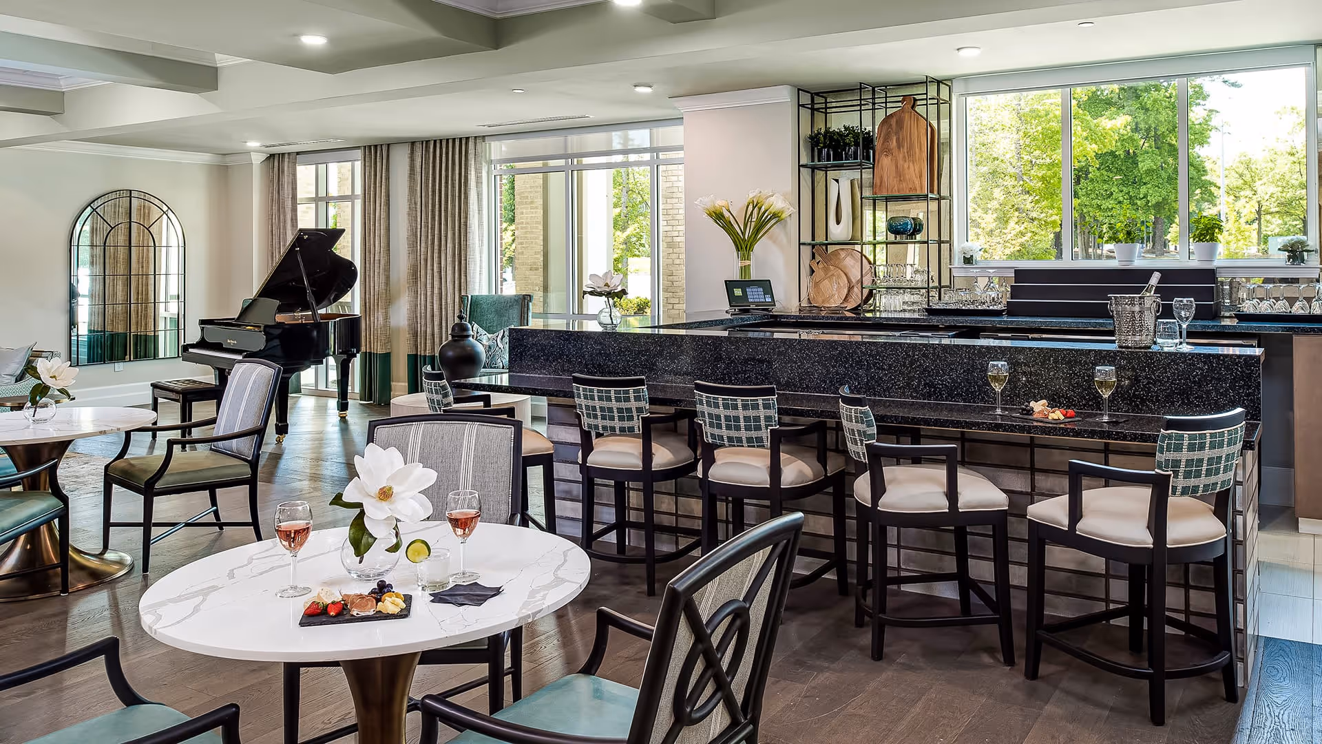 A bright and elegant common area with a black grand piano, several tables with chairs, and a bar counter with high stools. The tables have glasses of wine and small plates with snacks, along with white flower centerpieces. Large windows let in natural light and show green trees outside. The room has a modern and cozy atmosphere.