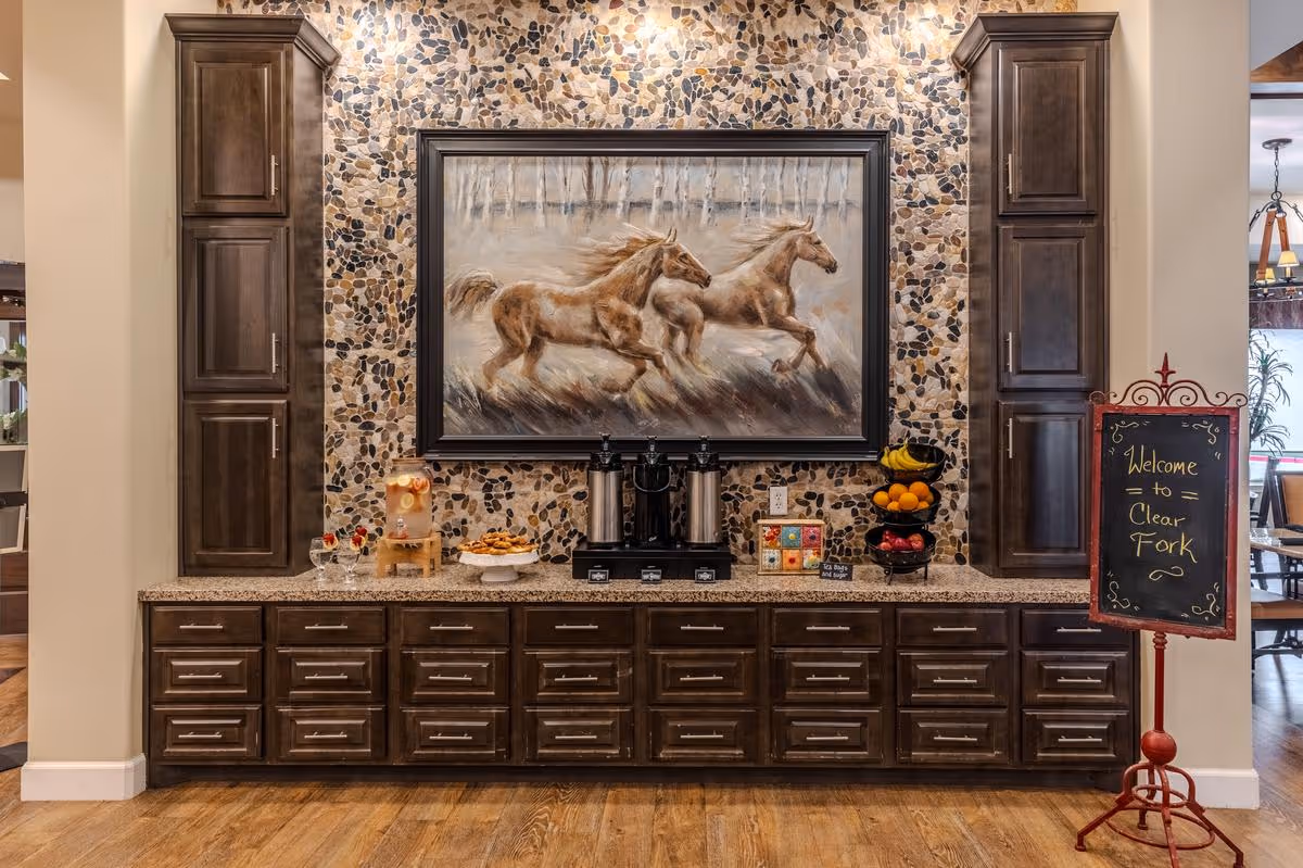 A countertop area with dark wooden cabinets and drawers, featuring a large framed painting of two running horses on a stone mosaic wall. On the countertop, there are beverage dispensers, a plate of cookies, glasses with drinks, a tiered fruit basket, and a small chalkboard sign that reads 'Welcome to Clear Fork'.