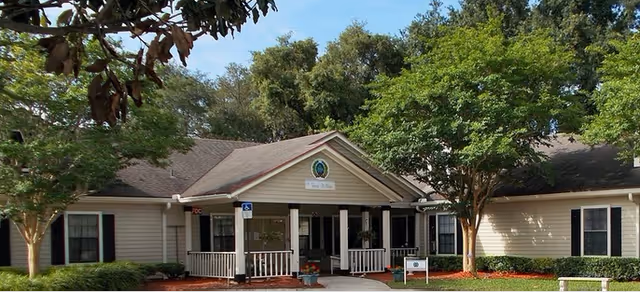 Front exterior of a single-story senior living facility with a covered entrance, white porch railing, and surrounding trees.