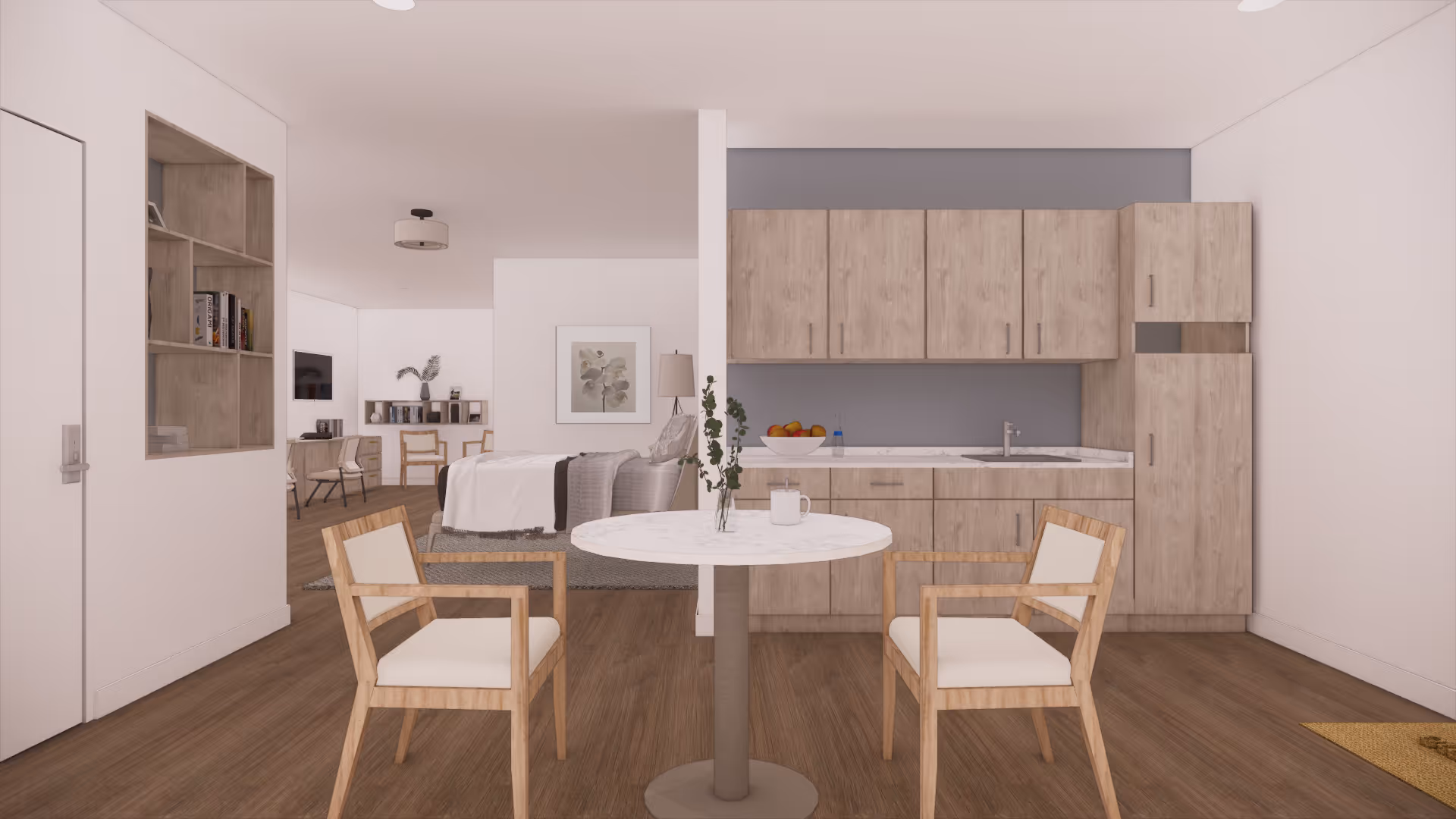 A modern assisted living apartment interior featuring a small round dining table with two wooden chairs in the foreground. Behind the table is a kitchenette with light wood cabinets, a sink, and a bowl of fruit. In the background, there is a bed with white bedding, a floor lamp, a framed botanical print on the wall, and a living area with a TV and additional seating. The flooring is wood, and the walls are painted white.
