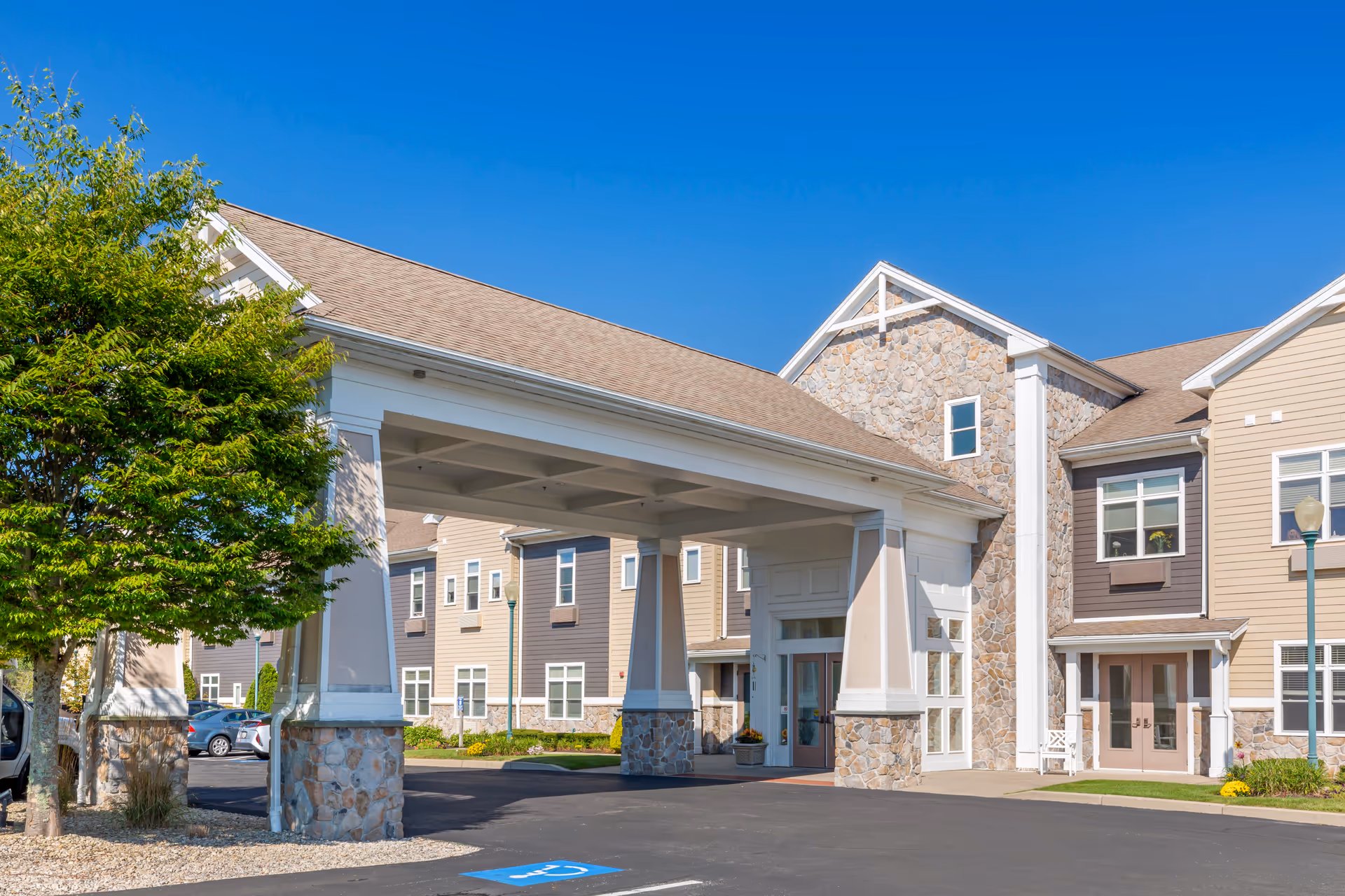 Exterior view of Brookdale Centre of New England showing the entrance with a covered drop-off area supported by stone and white pillars, beige and brown siding, multiple windows, a tree on the left, and a clear blue sky.