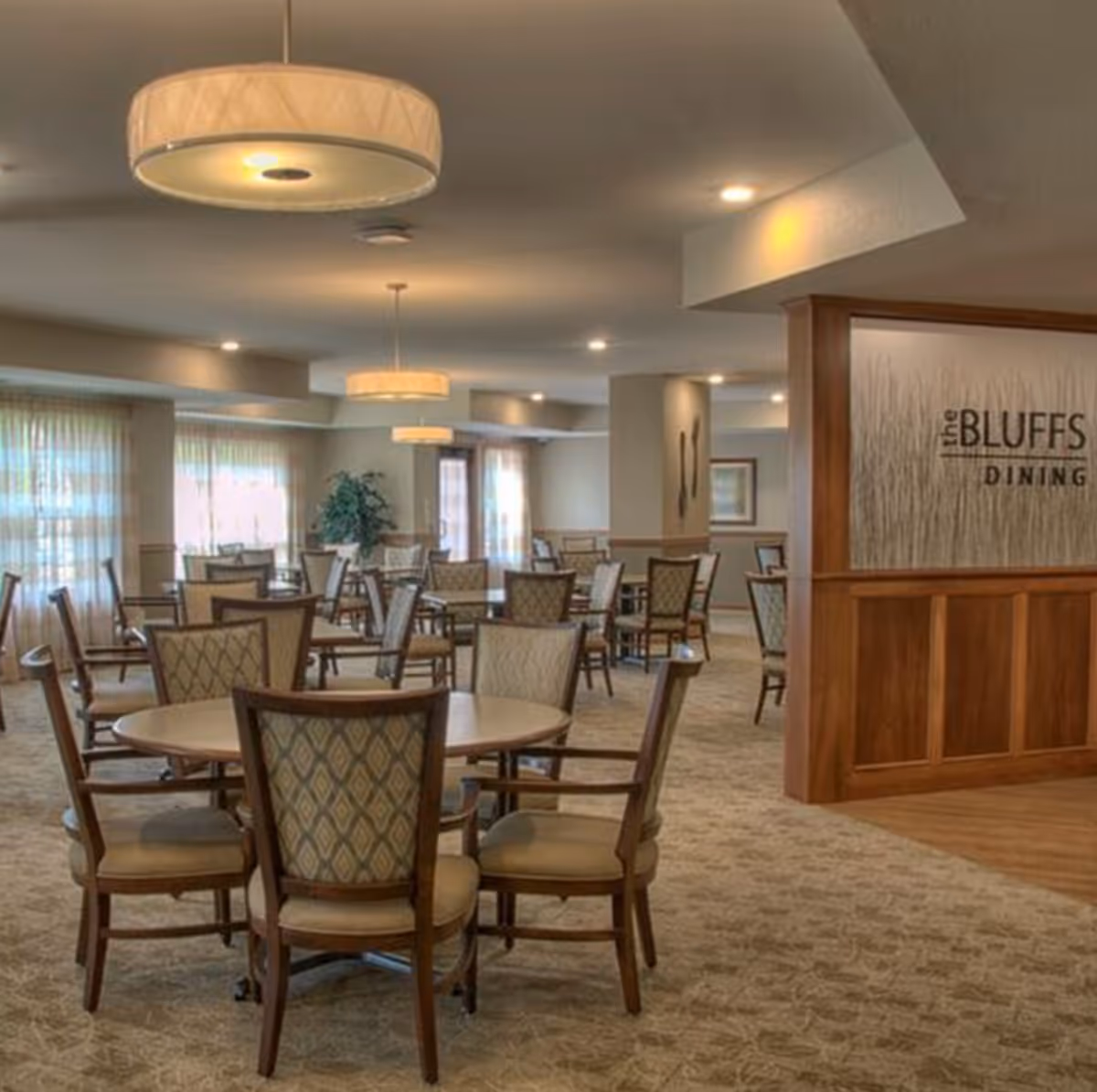 A spacious dining room with multiple round tables and cushioned chairs arranged neatly. The room is well-lit with large windows covered by sheer curtains and ceiling lights. A wooden partition on the right side displays a sign that reads 'the BLUFFS DINING'.