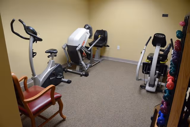 A small exercise room with beige walls and carpeted floor containing three pieces of exercise equipment including a stationary bike, a rowing machine, and a recumbent bike. There is a wooden chair with red cushioning in the corner and a rack of colorful dumbbells on the right side.