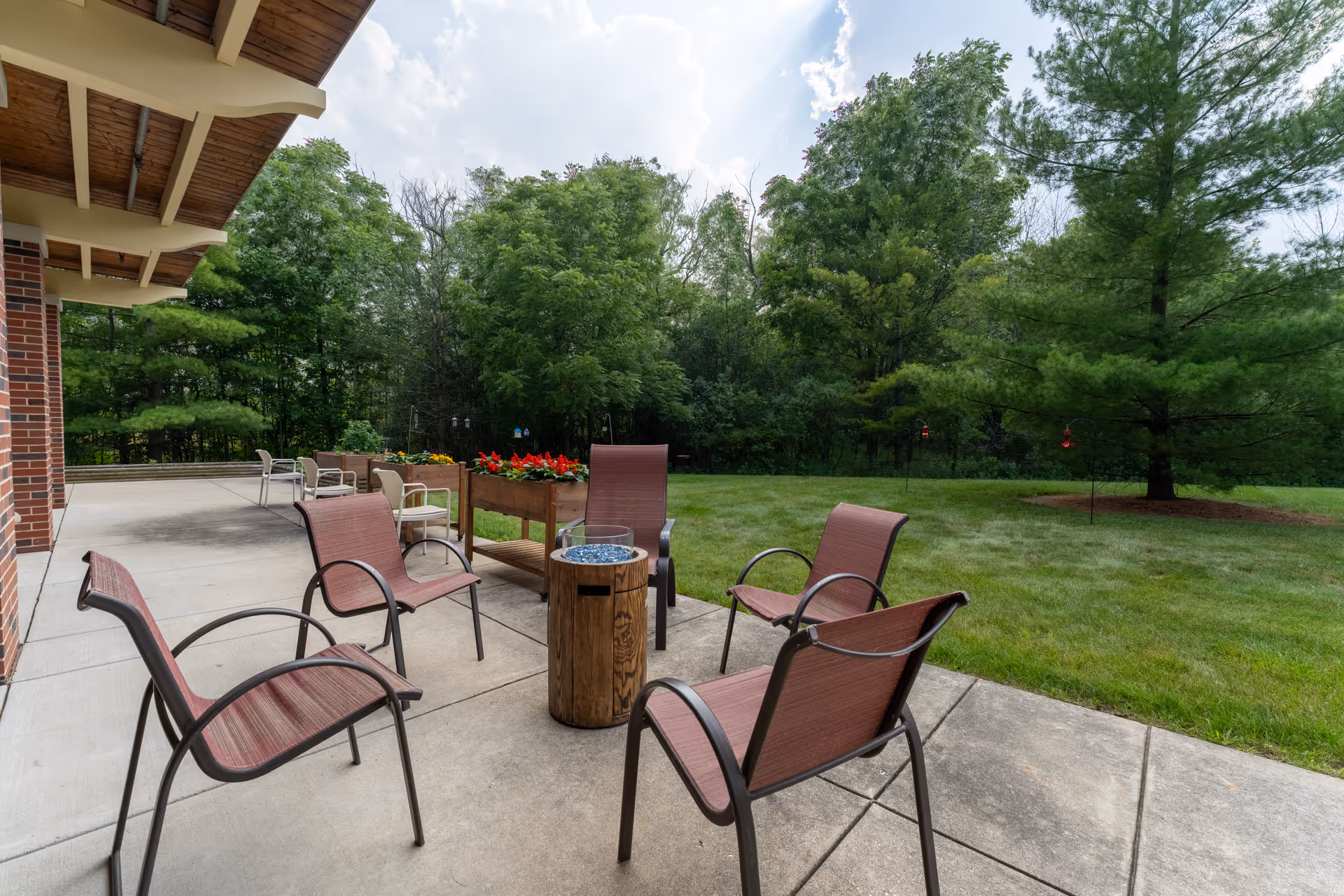 Outdoor patio area with five brown metal chairs arranged around a small wooden fire pit table. The patio is adjacent to a brick building and overlooks a grassy lawn with trees and flower planters in the background under a partly cloudy sky.