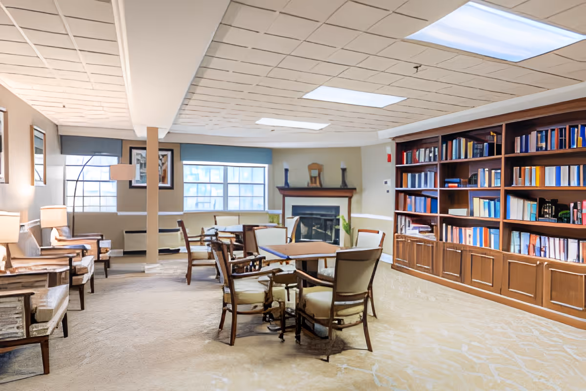 A cozy common area with several wooden chairs and tables arranged in the center. There is a large bookshelf filled with books along the right wall, and a fireplace with decorative items on the mantel in the background. The room has large windows with blue valances, beige walls, and carpeted flooring. Soft lighting is provided by lamps and ceiling lights.