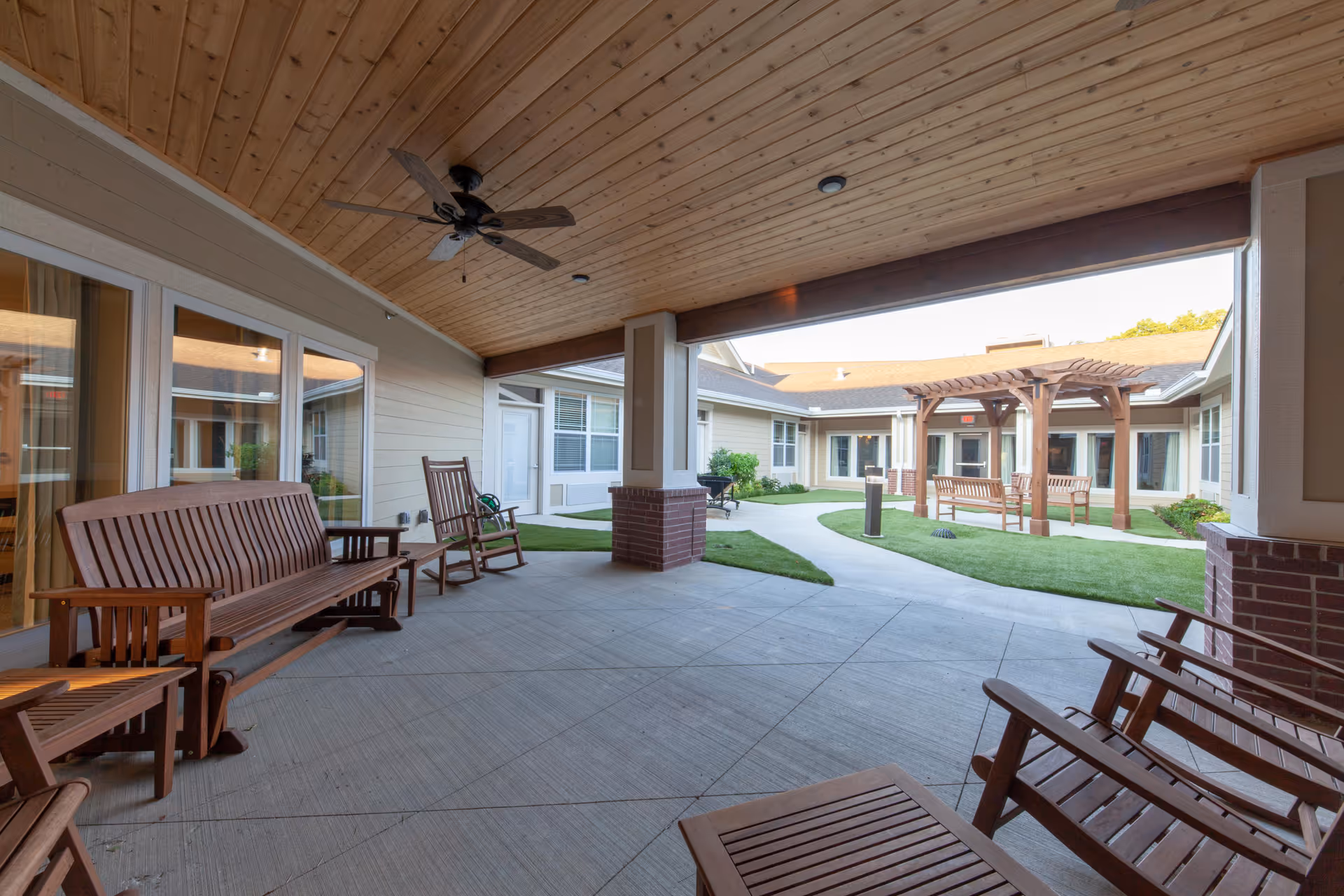 Covered outdoor patio area with wooden benches and rocking chairs, ceiling fan, and a view of a courtyard with a pergola and additional benches surrounded by grass and pathways at Cross Creek at Lee's Summit Memory Care & Assisted Living.