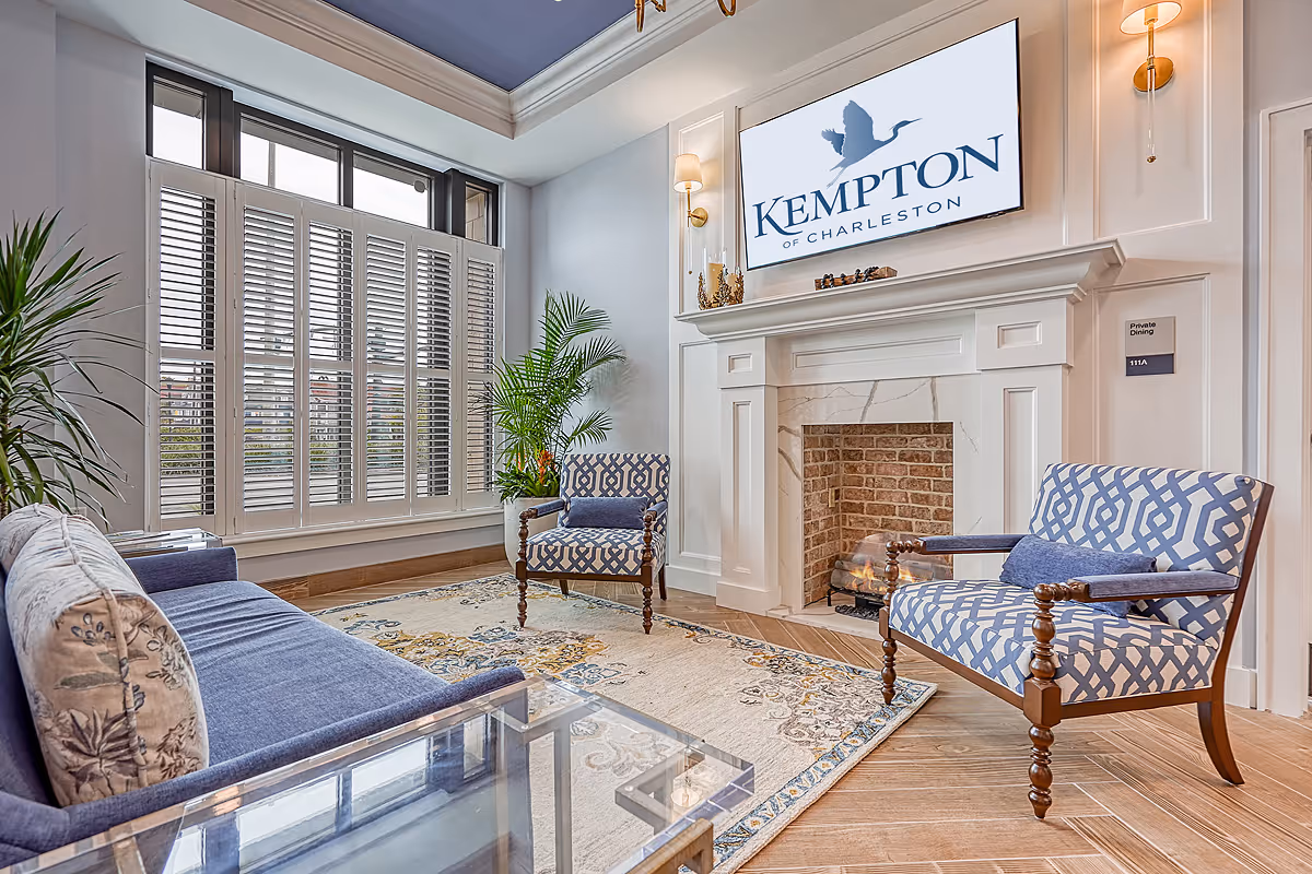 A bright and elegant living room area with a blue and white color scheme. The room features a blue sofa with patterned cushions, two blue and white patterned armchairs, a glass coffee table, a decorative rug, and a fireplace with a brick interior. Above the fireplace is a large sign that reads 'Kempton of Charleston'. Large windows with white shutters allow natural light to fill the space, and there are green plants adding a touch of nature to the room.