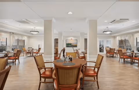 A spacious dining room with multiple round wooden tables and cushioned chairs arranged neatly. The room has large windows with sheer curtains allowing natural light to fill the space. The walls are light-colored with decorative molding, and ceiling lights provide additional illumination. A framed picture hangs on the far wall.