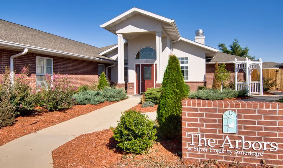 Exterior view of a senior living facility named The Arbors at Silver Creek by Americare, featuring a brick building with a red door, a walkway leading to the entrance, landscaped bushes, and a clear blue sky.