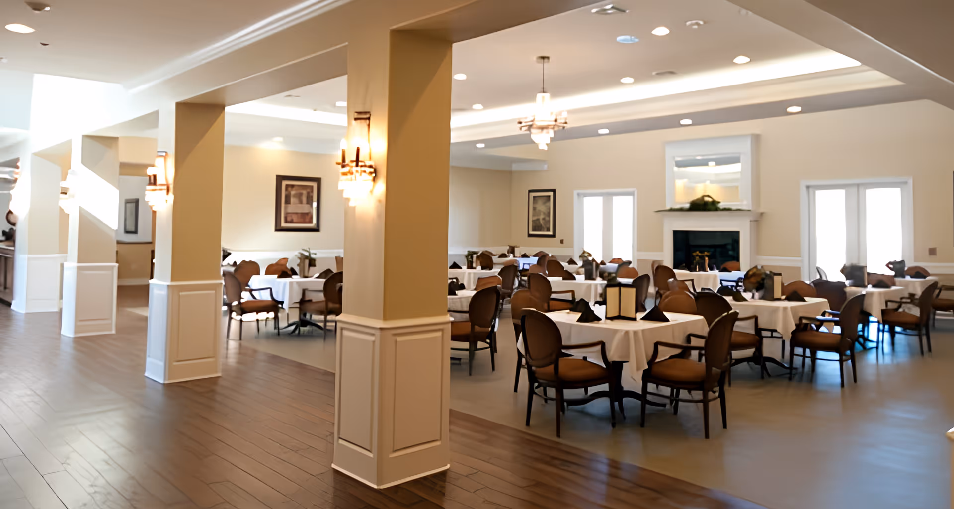 A spacious dining room in Pelican Bay Assisted Living Community with multiple round tables covered with white tablecloths, each surrounded by brown cushioned chairs. The room features beige walls, a fireplace with a mantel, framed artwork, and ceiling lights including chandeliers. The floor is a combination of wood and tile, and there are large windows allowing natural light to enter.