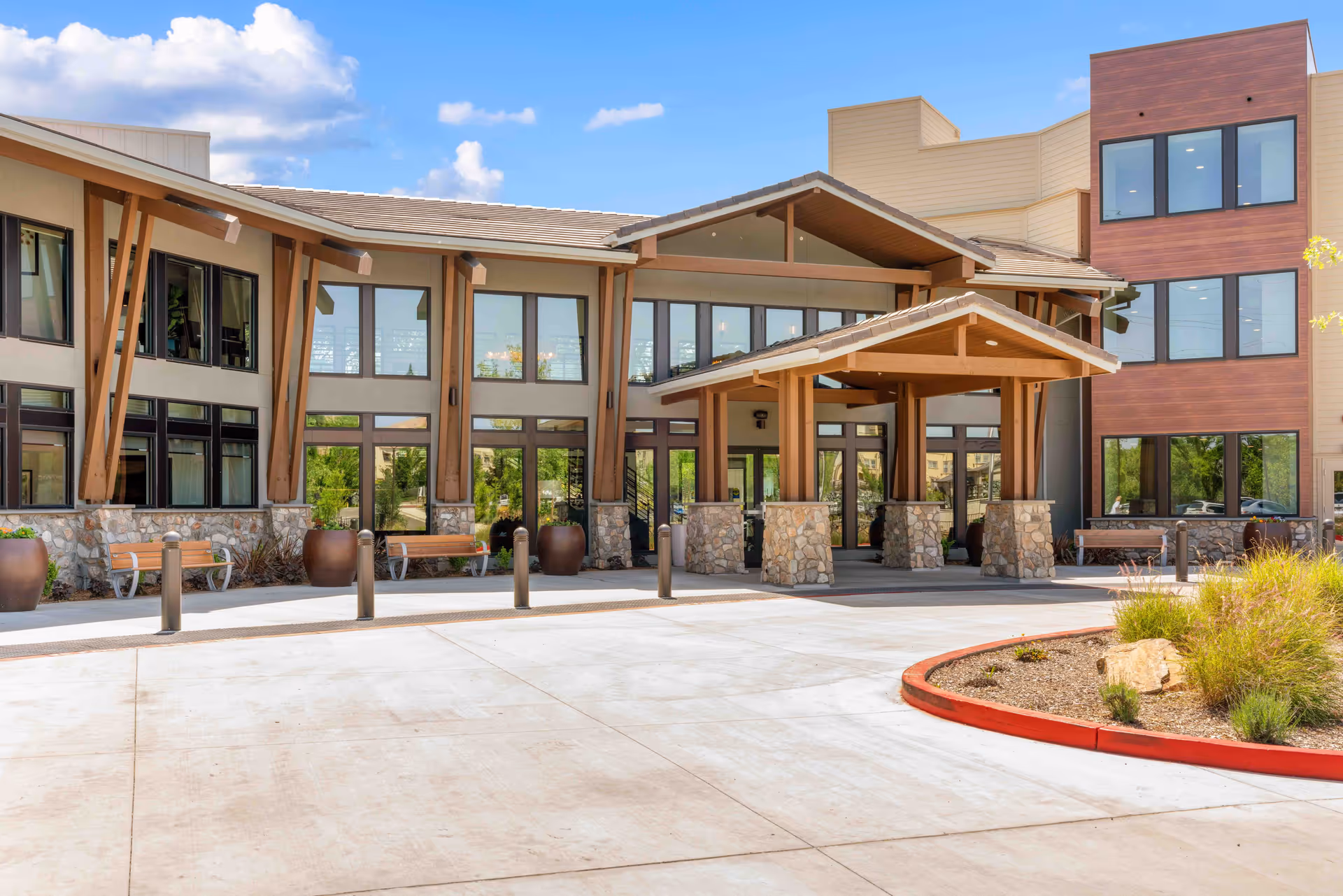 Exterior view of a modern senior living facility building with large windows, stone pillars, wooden beams, benches, and landscaped areas under a blue sky with some clouds.