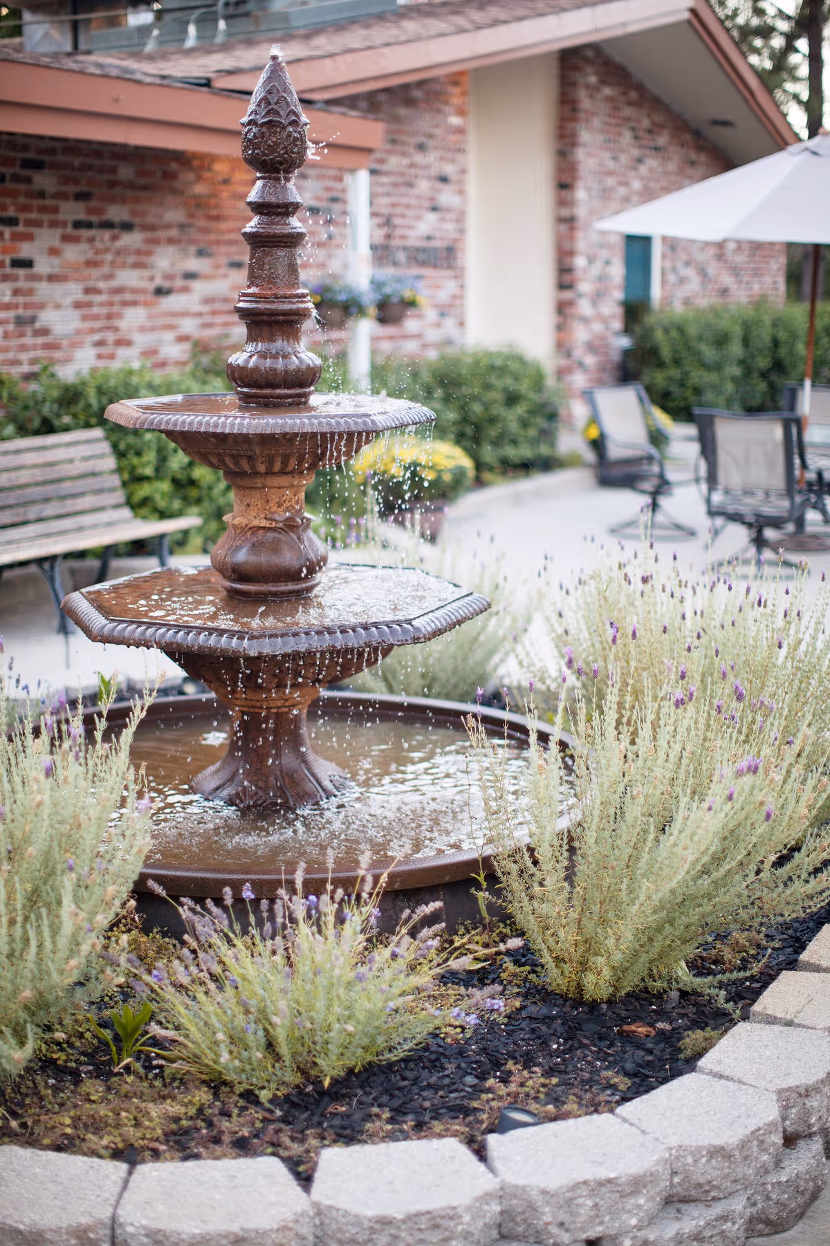 A decorative two-tiered water fountain surrounded by lavender plants and other greenery in a circular stone planter. In the background, there is a brick building, a wooden bench, patio chairs, and a white umbrella.