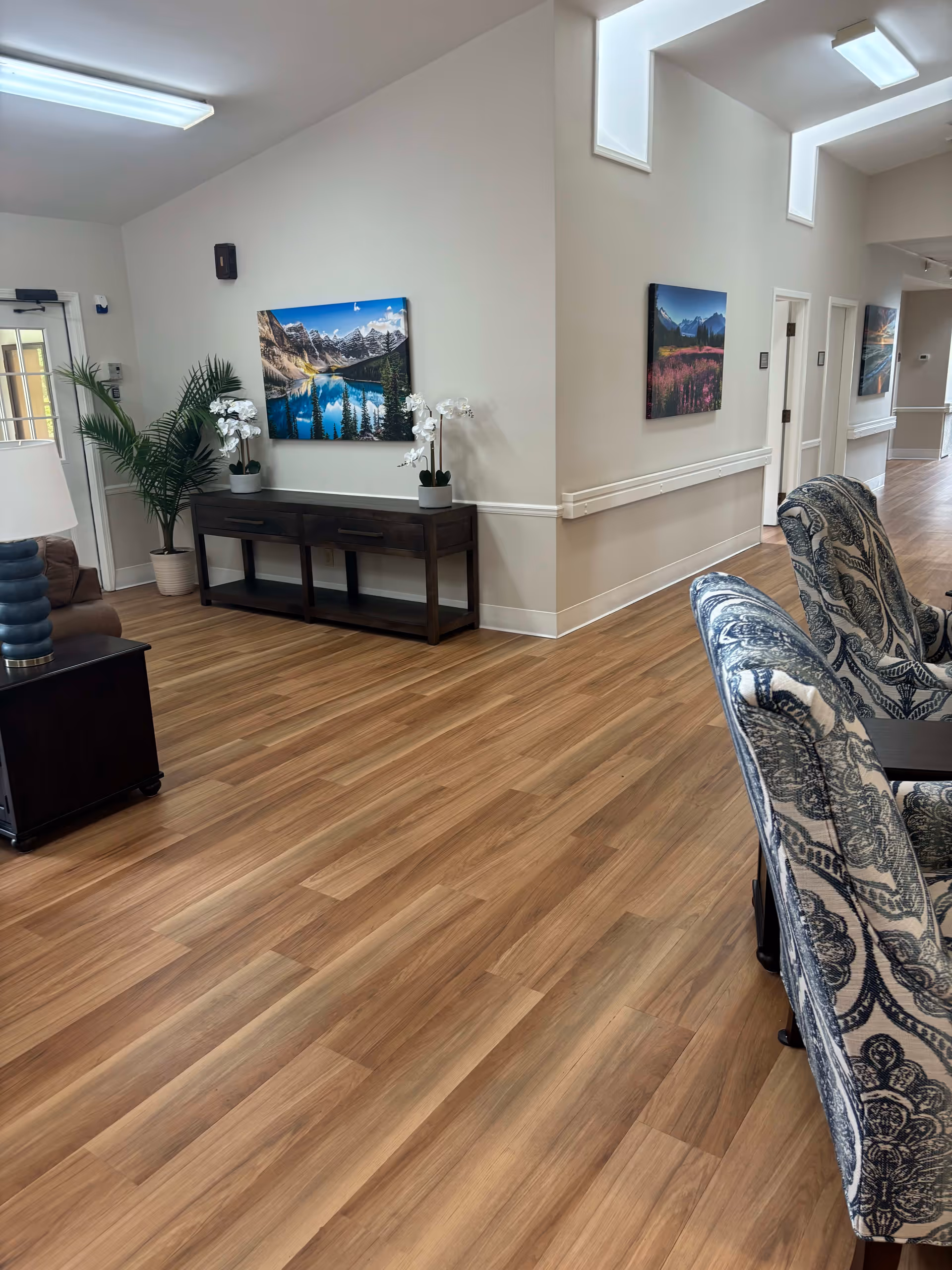 Interior hallway of South Knoxville Senior Living with wooden flooring, patterned armchairs, a dark wooden console table with white orchids, potted plants, and landscape paintings on the walls.