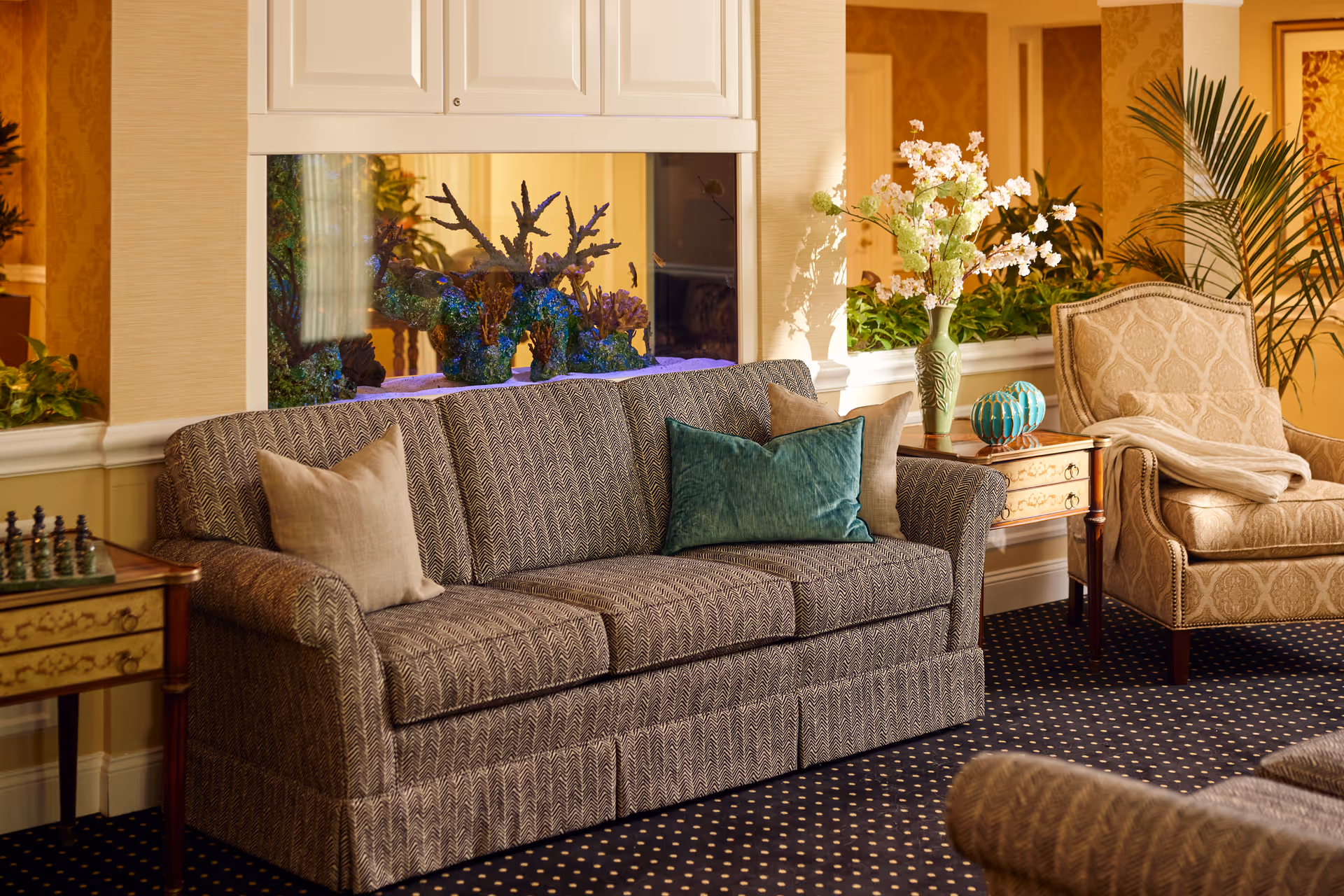 A cozy living room area featuring a patterned sofa with three cushions, a beige armchair with a throw blanket, two wooden side tables with decorative items including a vase with flowers and a small sculpture, and a built-in aquarium with coral decorations behind the sofa. The room has warm lighting, patterned carpet, and plants adding to the inviting atmosphere.