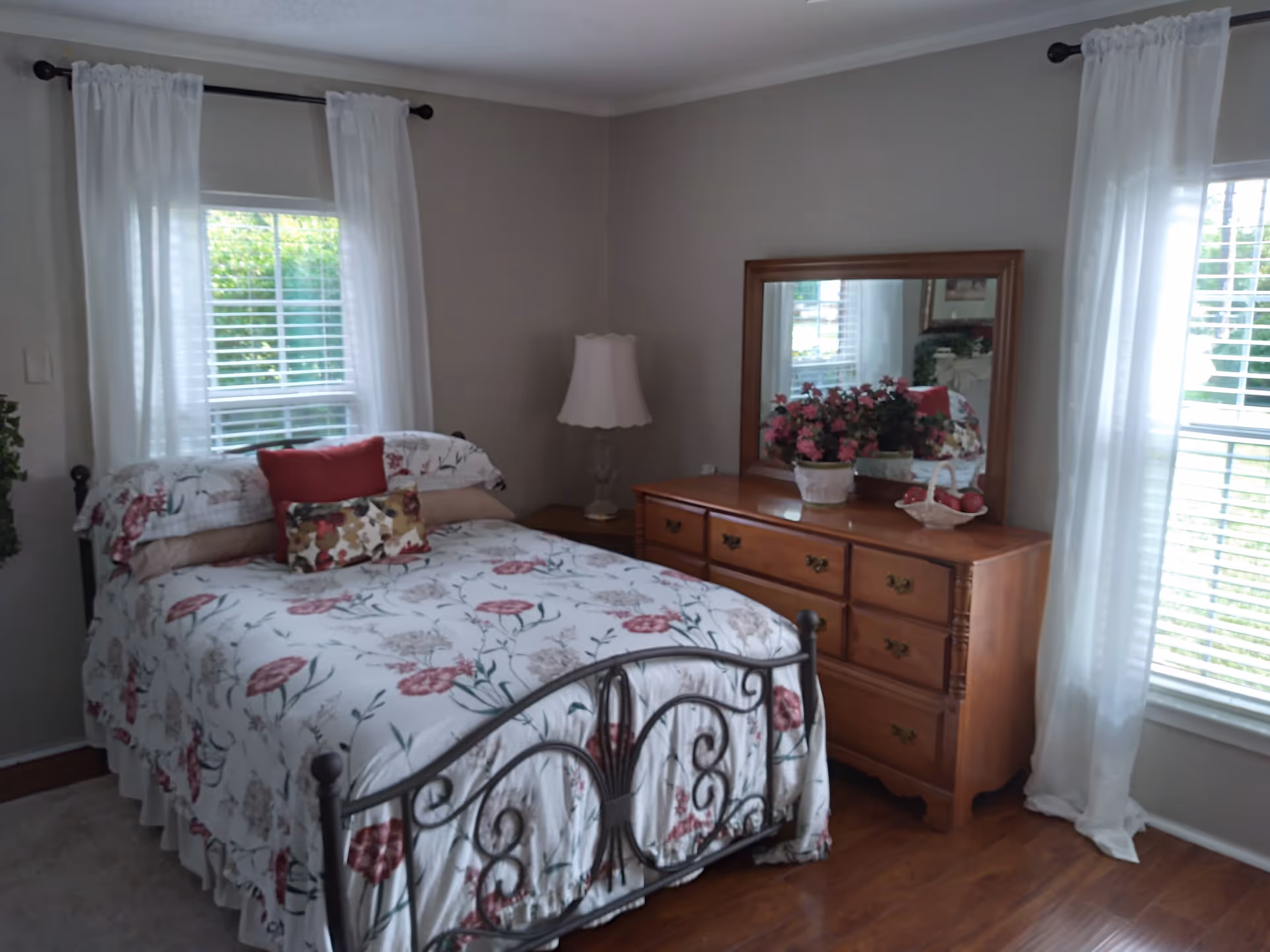 A cozy bedroom with a floral patterned bedspread on a metal bed frame, two windows with white curtains, a wooden dresser with a large mirror, a table lamp, and a vase of pink flowers on the dresser.