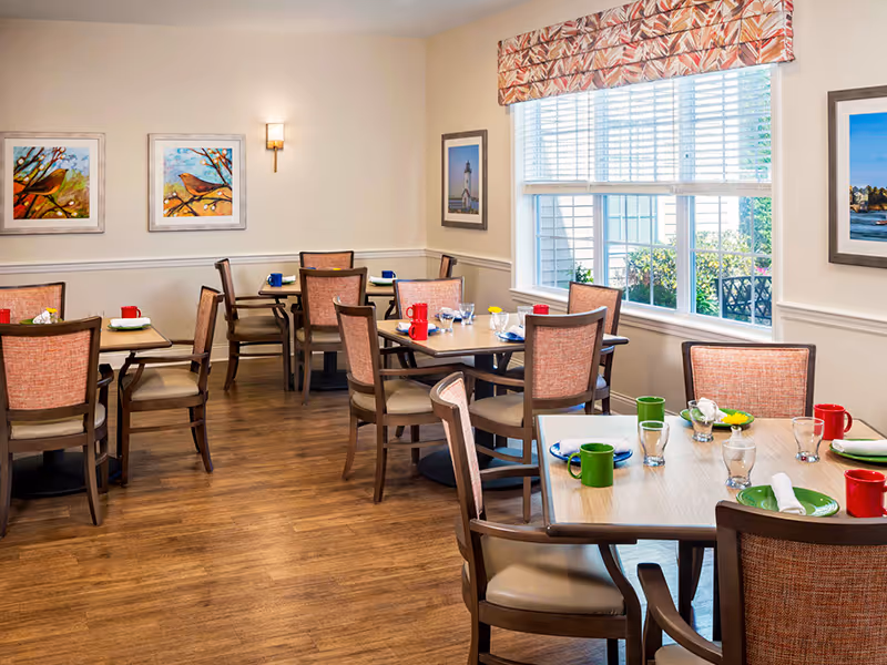 Dining room with wooden tables and upholstered chairs set with colorful cups and place settings beside a large window.