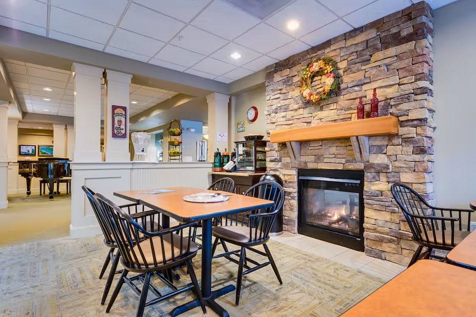 A cozy common area with a stone fireplace decorated with a floral wreath and red bottles on the mantle. In front of the fireplace are wooden tables with black chairs. The room has a carpeted floor and a ceiling with recessed lighting. In the background, there is a water dispenser and a piano visible in an adjacent room.