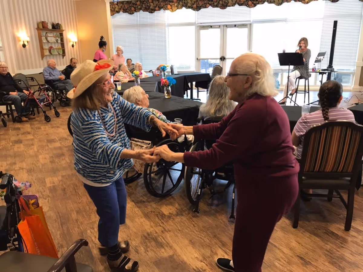 Two elderly women hold hands and dance in a communal activity room while other residents watch and a musician performs.