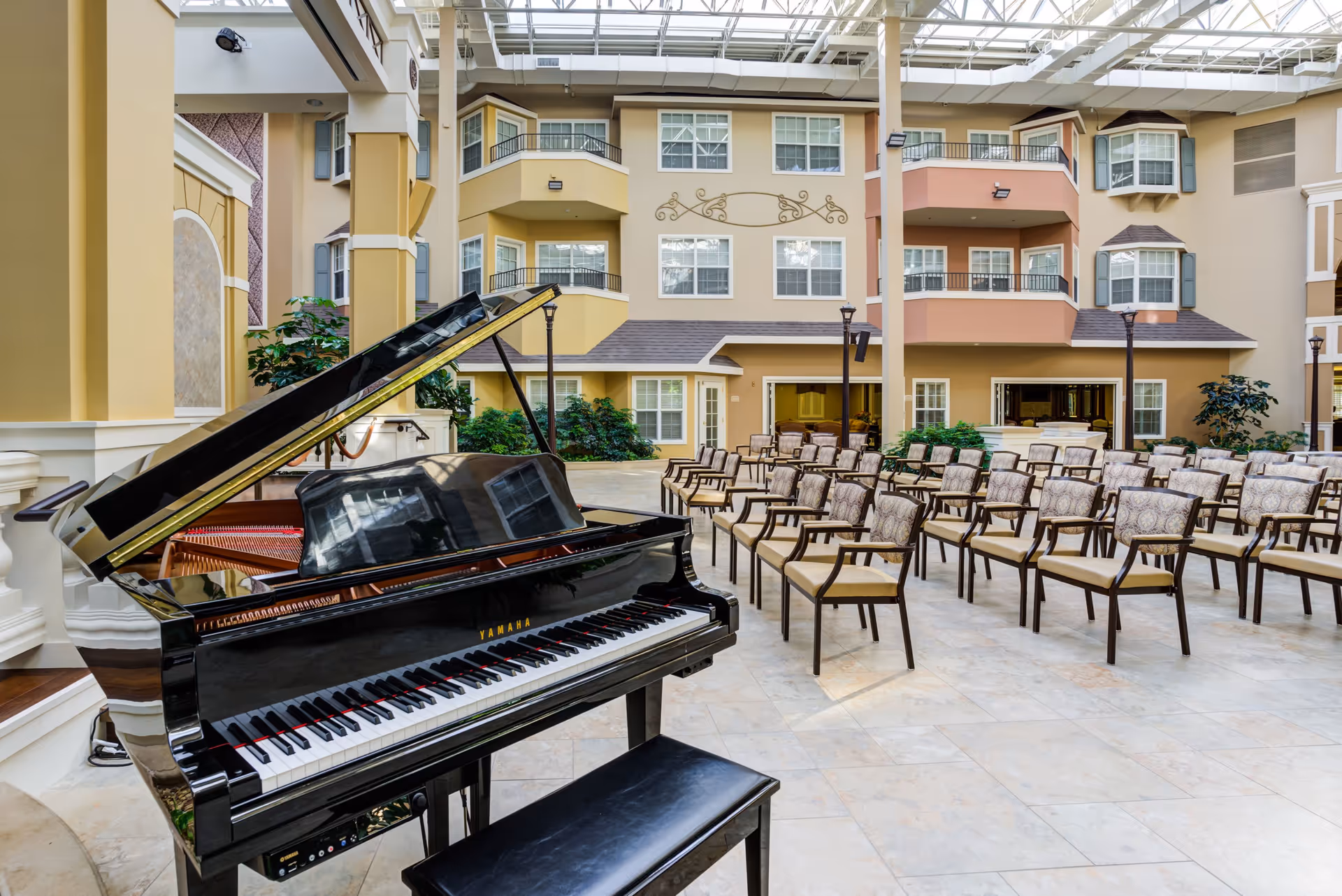 Indoor atrium area with a black Yamaha grand piano in the foreground and rows of cushioned chairs arranged for an audience. The space is surrounded by multi-story residential-style walls with windows and balconies, and a glass ceiling allowing natural light to fill the area.