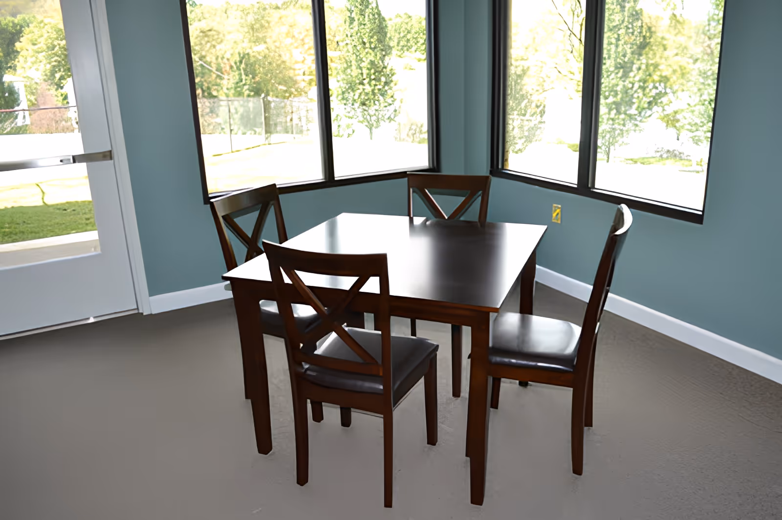 A small dining area with a square wooden table and four matching wooden chairs with cushioned seats. The room has large windows letting in natural light and a door leading outside. The walls are painted a soft blue color and the floor is carpeted.