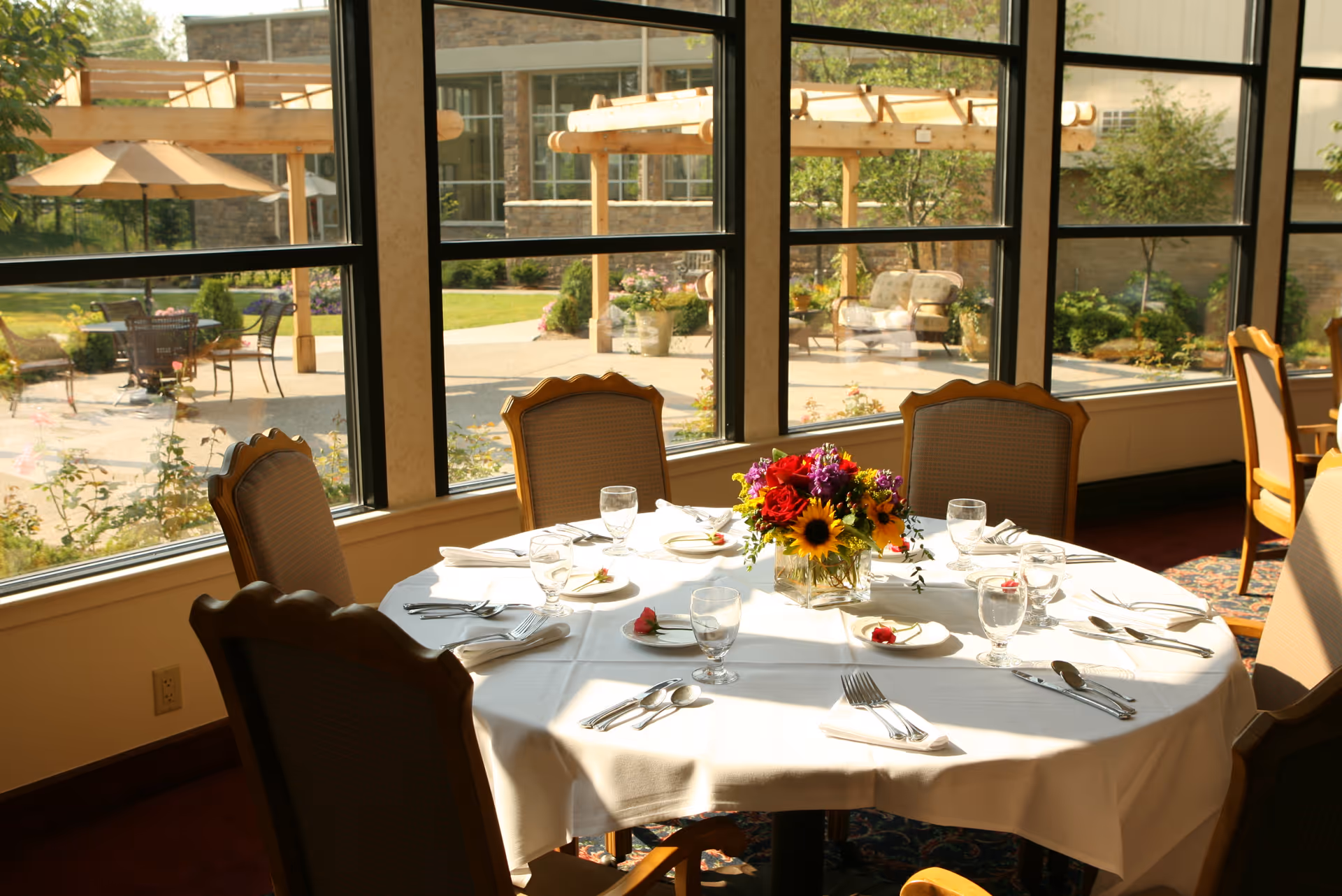 A round dining table set for six with white tablecloth, glassware, silverware, and a colorful floral centerpiece, situated next to large windows overlooking an outdoor patio with tables, chairs, umbrellas, and pergolas surrounded by greenery.