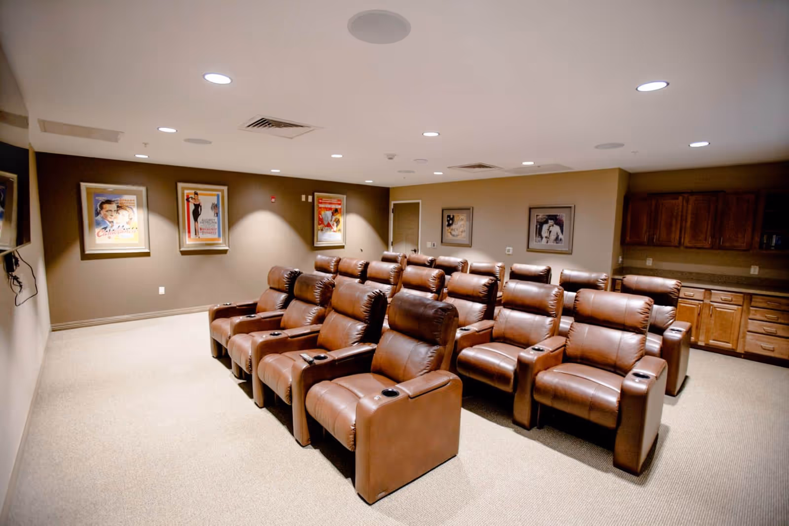 Rows of brown leather recliner chairs arranged theater-style in a carpeted media room with framed posters on the walls and cabinets at the back.