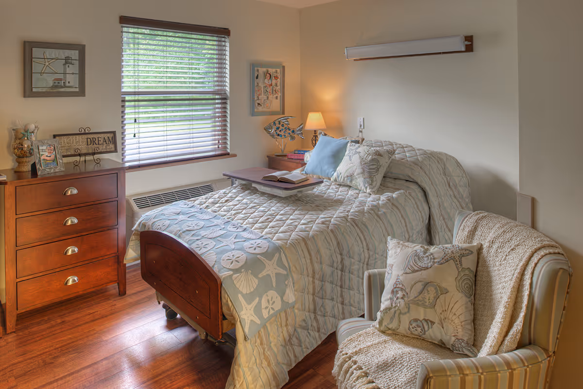A cozy bedroom in a senior living facility with a single bed covered in a quilted bedspread featuring seashell and starfish patterns. There is a wooden dresser with decorative items and a framed picture on it, a window with blinds letting in natural light, a bedside table with a lamp and books, and a comfortable armchair with a throw blanket and a seashell-patterned pillow.