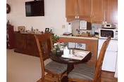 Interior view of a retirement community living space featuring a small round dining table with two wooden chairs, a flower centerpiece, and place settings. In the background, there is a wooden dresser with decorative items and a wall-mounted television. A kitchenette area with cabinets, a microwave, and a small refrigerator is also visible.