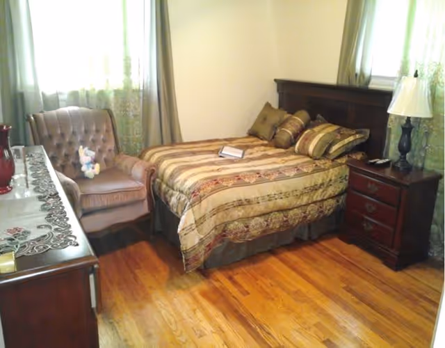 A cozy bedroom with a wooden floor, a bed with a striped and patterned comforter and several pillows, a wooden nightstand with a lamp and a phone, a cushioned armchair with a stuffed toy on it, and a dresser with decorative items. Light filters through windows with curtains.