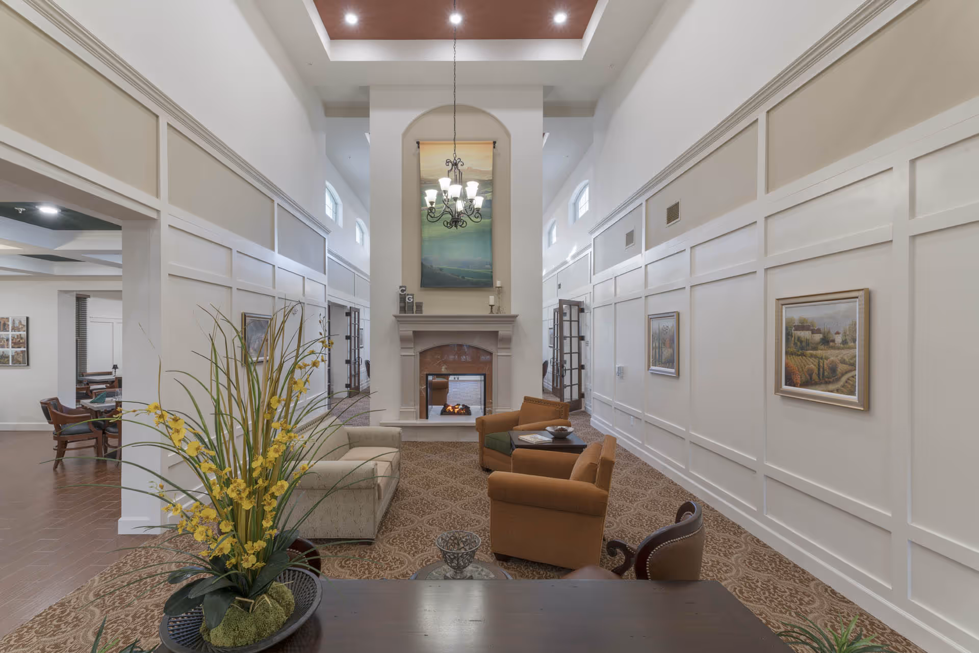 A spacious and elegant living room area with high ceilings, a central fireplace, and a chandelier hanging above. The room features a beige sofa, two brown armchairs, a small table, and decorative paintings on the walls. There is a large floral arrangement on a wooden table in the foreground.