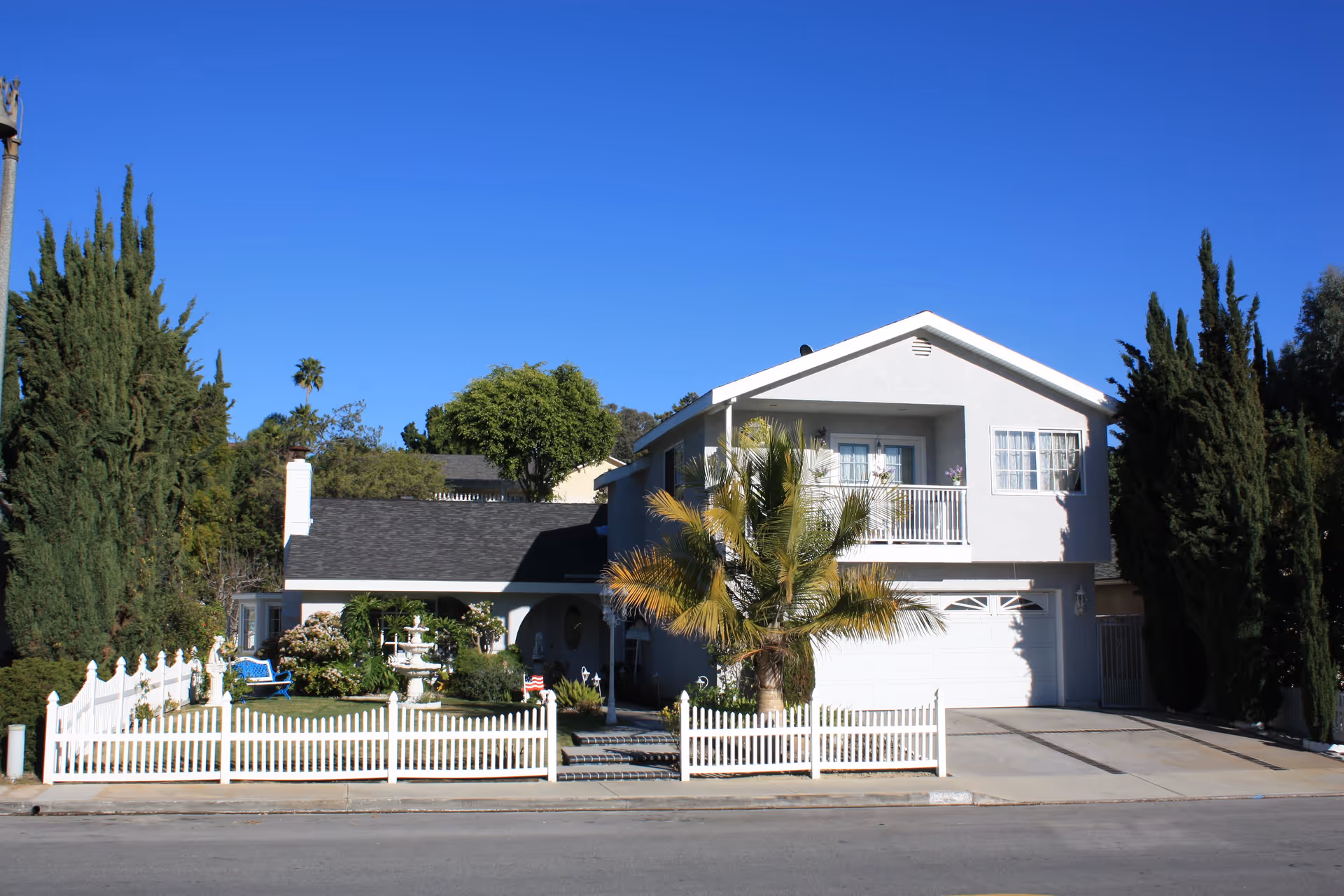 A two-story residential building with a white picket fence, a driveway, and a small garden with a fountain and palm tree in front. The house has a balcony on the upper floor and is surrounded by tall trees under a clear blue sky.