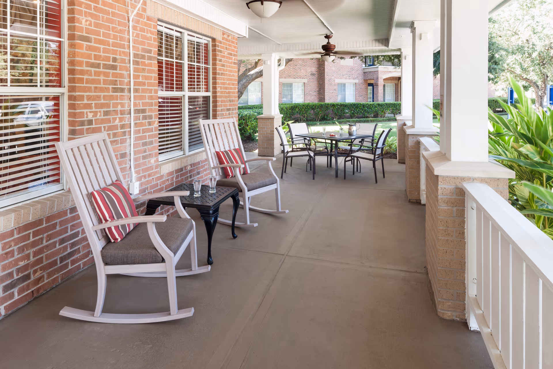 A covered outdoor porch area with two white rocking chairs with cushions and a small black table between them. Further down the porch is a round glass table with four chairs. The porch has brick walls, white columns, ceiling fans, and overlooks a green garden area with bushes and trees.