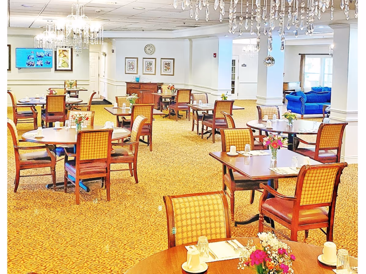 A bright and elegant dining room with multiple round and square wooden tables, each set with cups, glasses, and napkins. The chairs have wooden frames with patterned upholstery. Crystal chandeliers hang from the ceiling, and there are floral centerpieces on the tables. In the background, there is a blue couch and framed artwork on the walls.