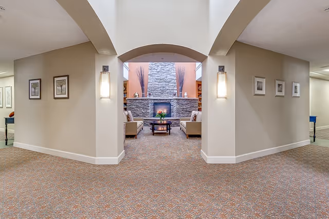 View through an arched hallway into a cozy sitting area with a stone fireplace, two beige armchairs, a coffee table with flowers, and built-in shelves on either side of the fireplace. The hallway walls are decorated with framed pictures and wall sconces, and the floor is carpeted with a patterned design.
