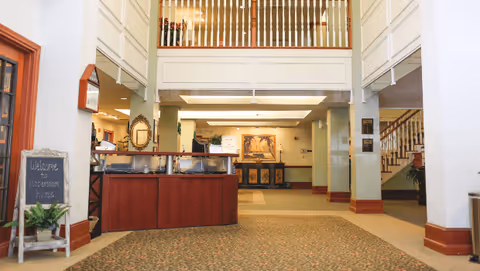 Interior view of a senior living facility lobby area with a reception desk in the center, a staircase on the right, and a welcoming sign on a small chalkboard on the left that reads 'Welcome to Habersham House'. The space has a high ceiling with a balcony railing above and warm lighting.