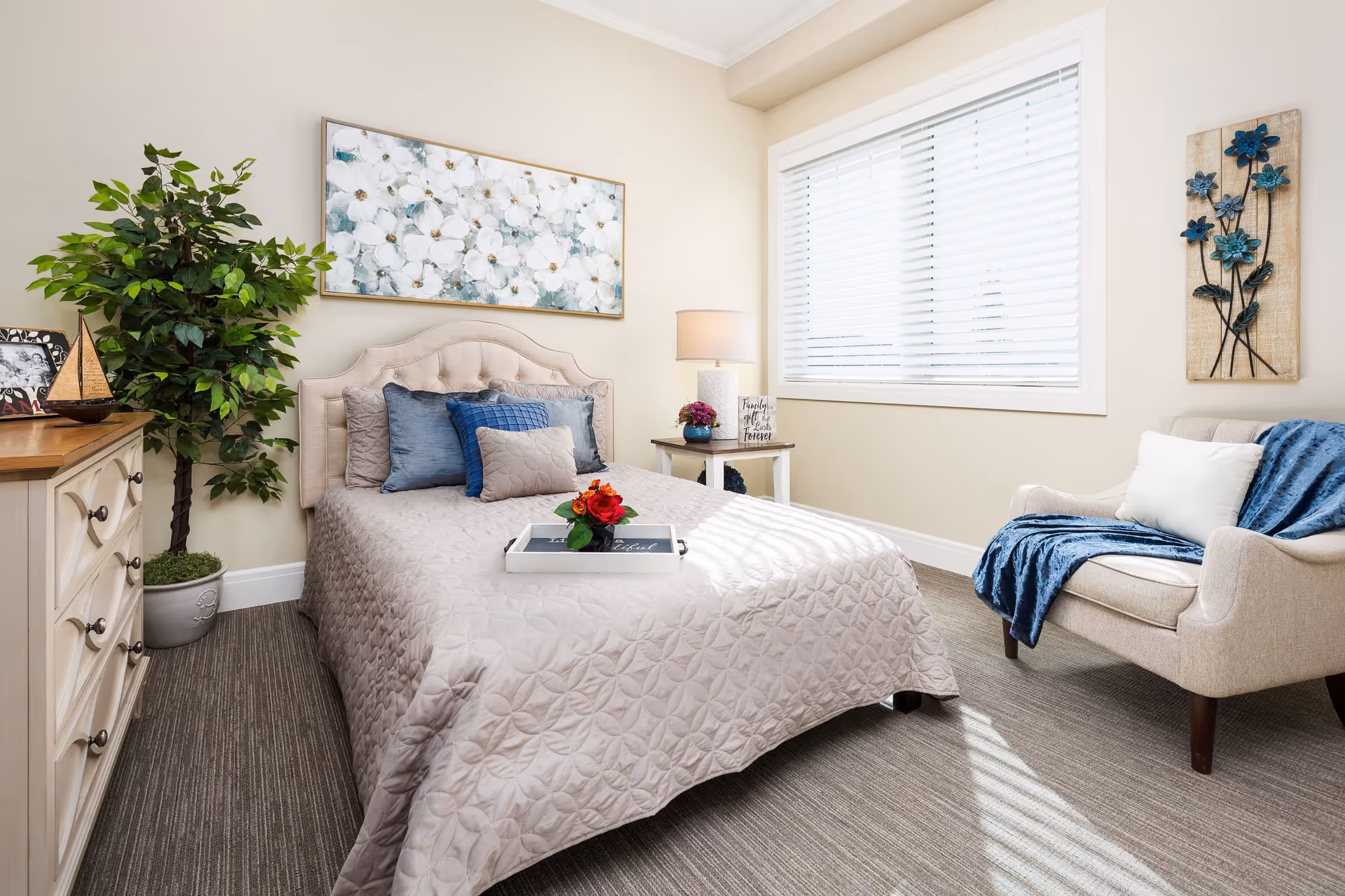 Sunlit, tastefully decorated bedroom featuring a bed with blue pillows, an armchair with a throw, a dresser, and a potted plant.