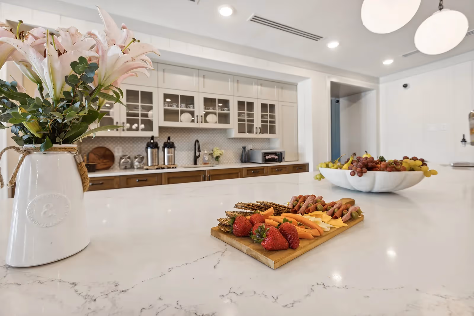 A modern kitchen with a white marble countertop featuring a wooden board with strawberries, cheese, crackers, and carrot sticks. A white vase with pink lilies and greenery is on the left side of the counter. In the background, there are white cabinets with glass doors, a sink, a microwave, and two coffee dispensers.