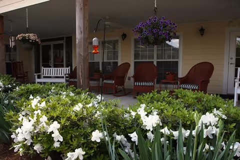 Covered patio area at Creekside Pines Retirement Community with red cushioned chairs, a white bench, hanging flower pots, and a hummingbird feeder, surrounded by green shrubs and white flowers.