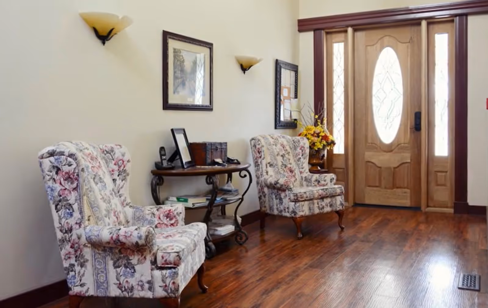 A cozy interior entryway with two floral upholstered armchairs, a wooden side table holding a phone, picture frame, and decorative box, wall-mounted lights, framed artwork, and a wooden front door with glass panels and decorative designs.