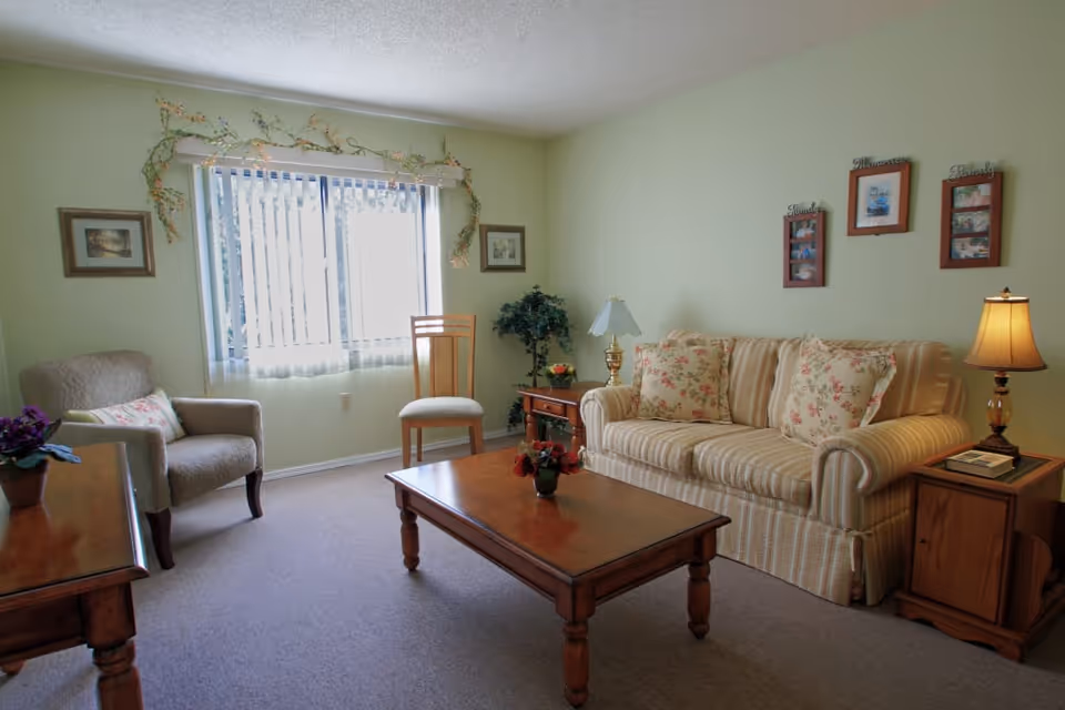 A cozy living room with a striped sofa adorned with floral cushions, a wooden coffee table with a small flower arrangement, an armchair with a decorative pillow, a wooden chair, and two side tables each with a lamp. The walls are light green with framed pictures and a window with vertical blinds letting in natural light.