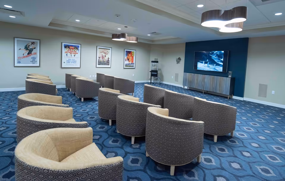 A cozy media room with multiple rows of curved armchairs facing a large flat-screen TV mounted on a dark blue accent wall. The room has a patterned blue carpet, framed vintage movie posters on the wall, and a popcorn machine in the corner.
