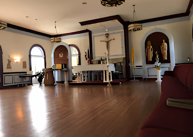 A spacious room with wooden flooring and arched windows. The room features religious statues and a large crucifix mounted on the wall. There is a long maroon cushioned bench along one side and a lectern near the windows. The room appears to be a chapel or prayer room within a senior living facility.