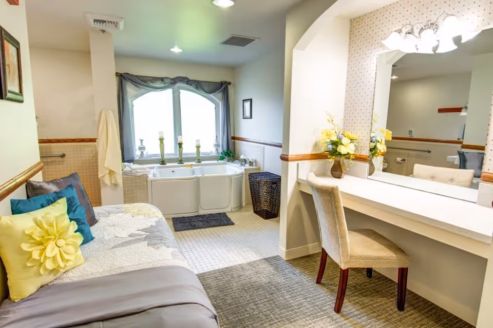 A bright and cozy room featuring a walk-in bathtub near a window with decorative candles, a cushioned bench with colorful pillows, and a vanity area with a large mirror, chair, and floral arrangement.