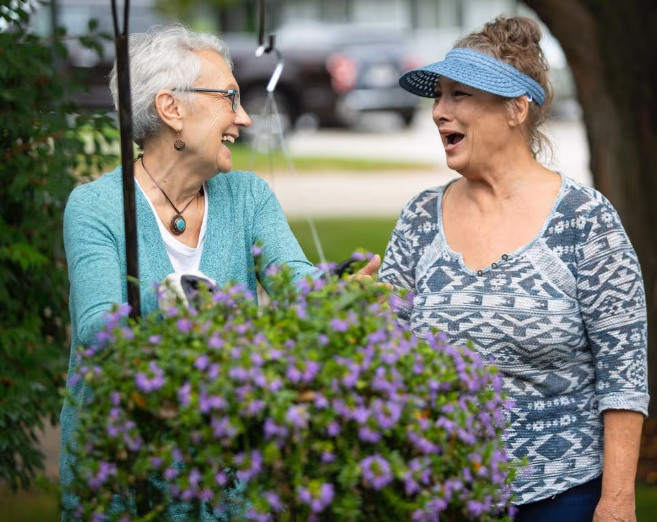 Two older women smiling and engaging in conversation outdoors near a hanging basket of purple flowers, with greenery and a blurred background of parked cars.
