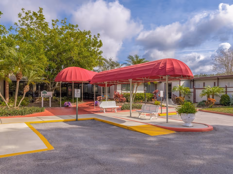 Entrance area of Woodlands Village Senior Living Community featuring two red fabric canopies over white benches, surrounded by landscaped greenery including palm trees and bushes, with a partly cloudy sky overhead.
